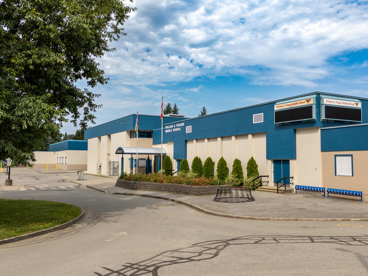 Exterior image of a middle school with a blue sky and some clouds. It shows the driveway entrance to the school main office and parking lot at the front of the school.